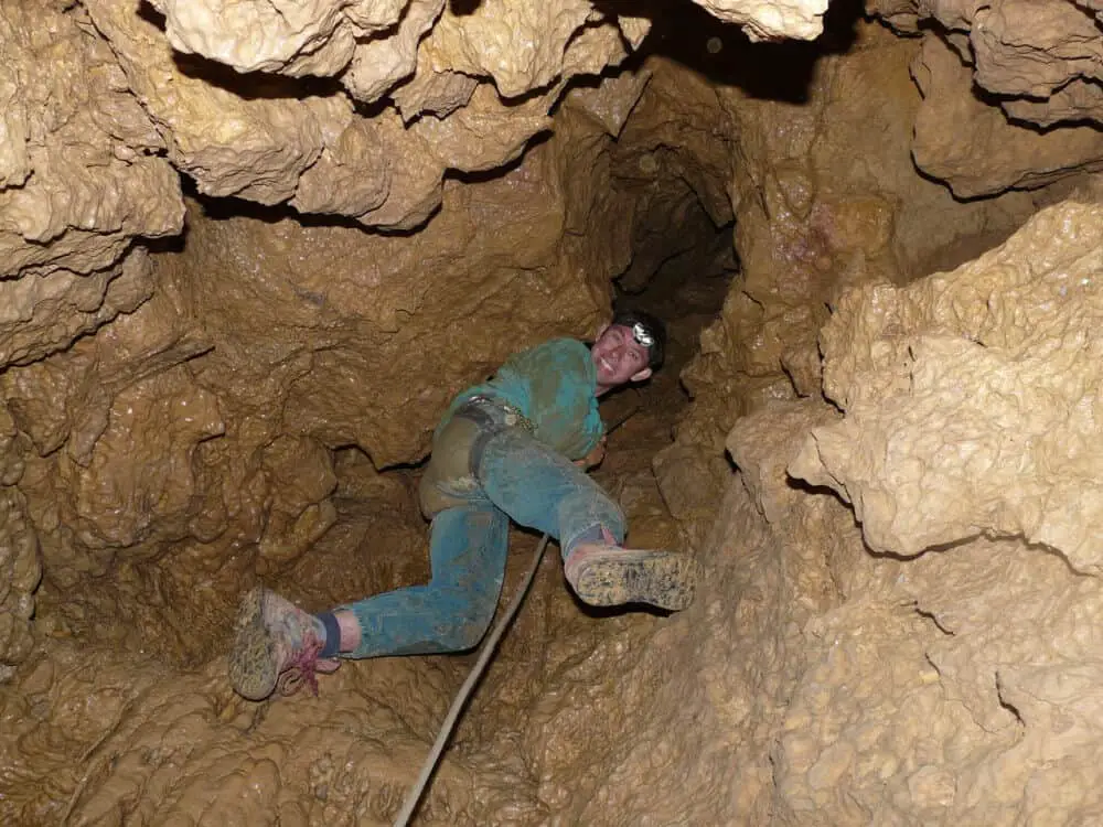 A smiling caver ascending a rope through mud and popcorn rock